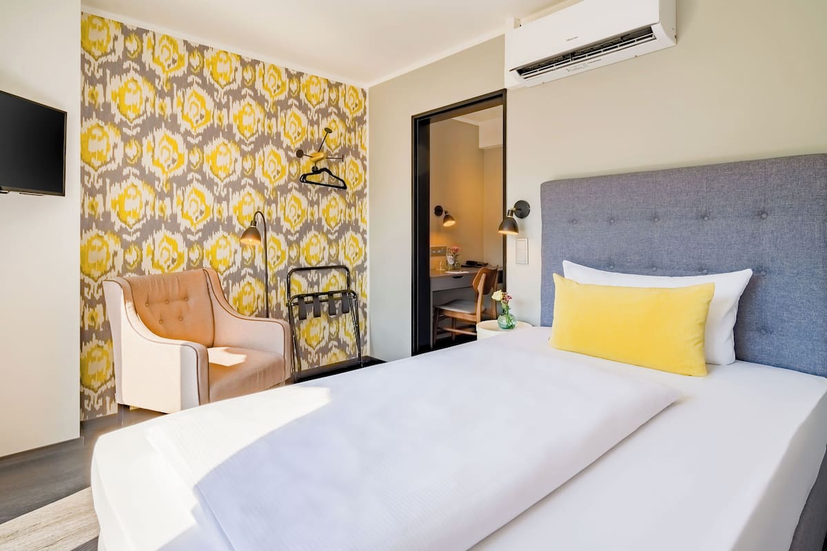 A hotel room with a bed, a chair, a desk, and a patterned wallpaper.
