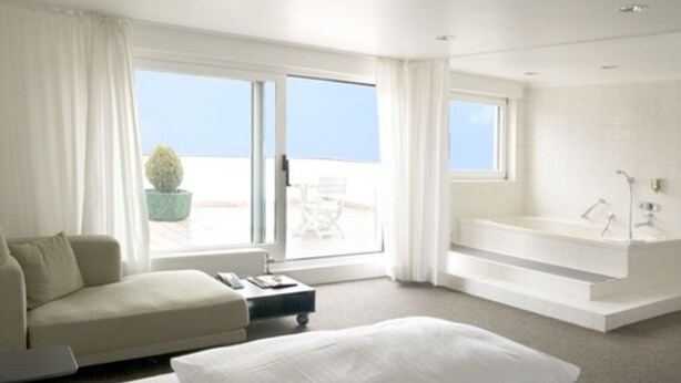 A modern hotel room with a large bed, a sofa, a small table, a bathtub, and a view of the sea. - rugs HOTEL Köln (Cologne)