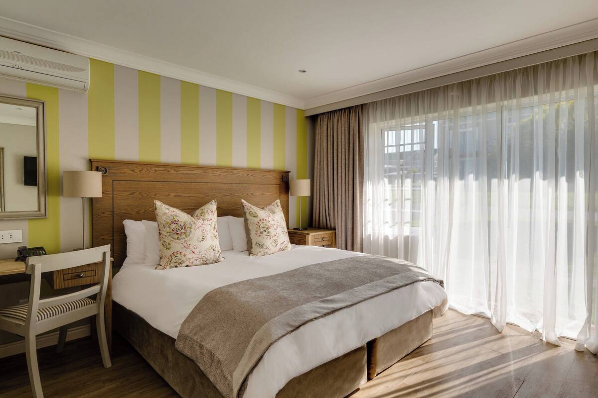 Een hotelkamer met een groot bed, een houten hoofdeinde, twee nachtkastjes met lampen, een stoel en een groot raam met dunne gordijnen.