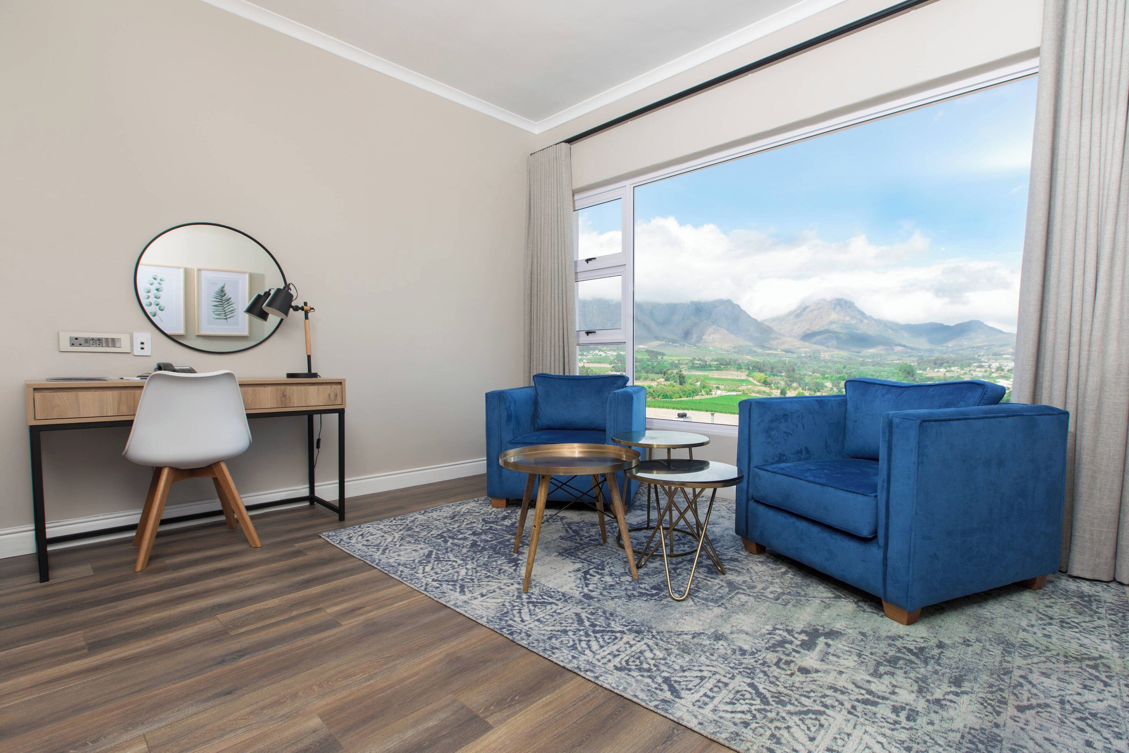 Une chambre d’hôtel avec un bureau, deux fauteuils bleus, un miroir rond et une vue sur les montagnes.