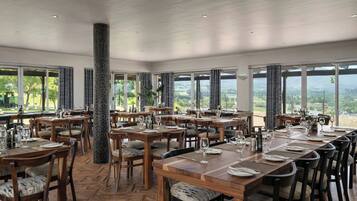 A spacious dining area with multiple tables set for dining, large windows offering a view of the outdoors, and a high ceiling with recessed lighting.
