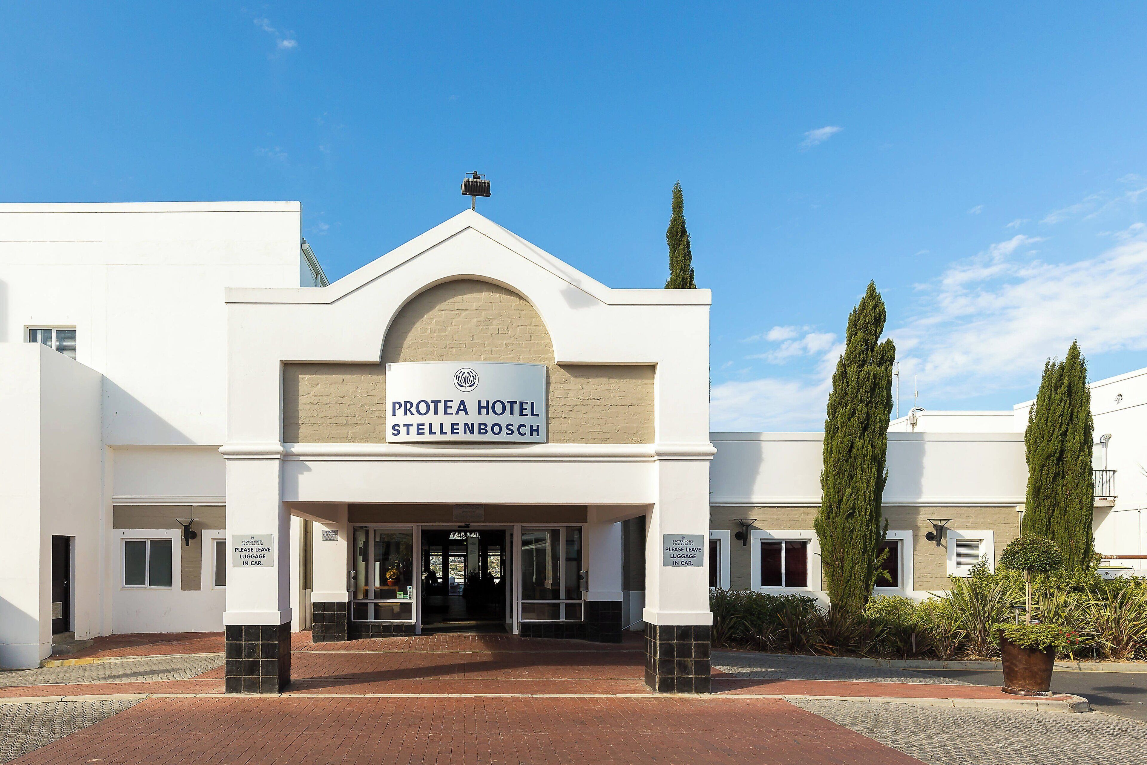 Exterior do Protea Hotel Stellenbosch, com entrada visível, placa indicando armazenamento de bagagem e fachada bem cuidada.