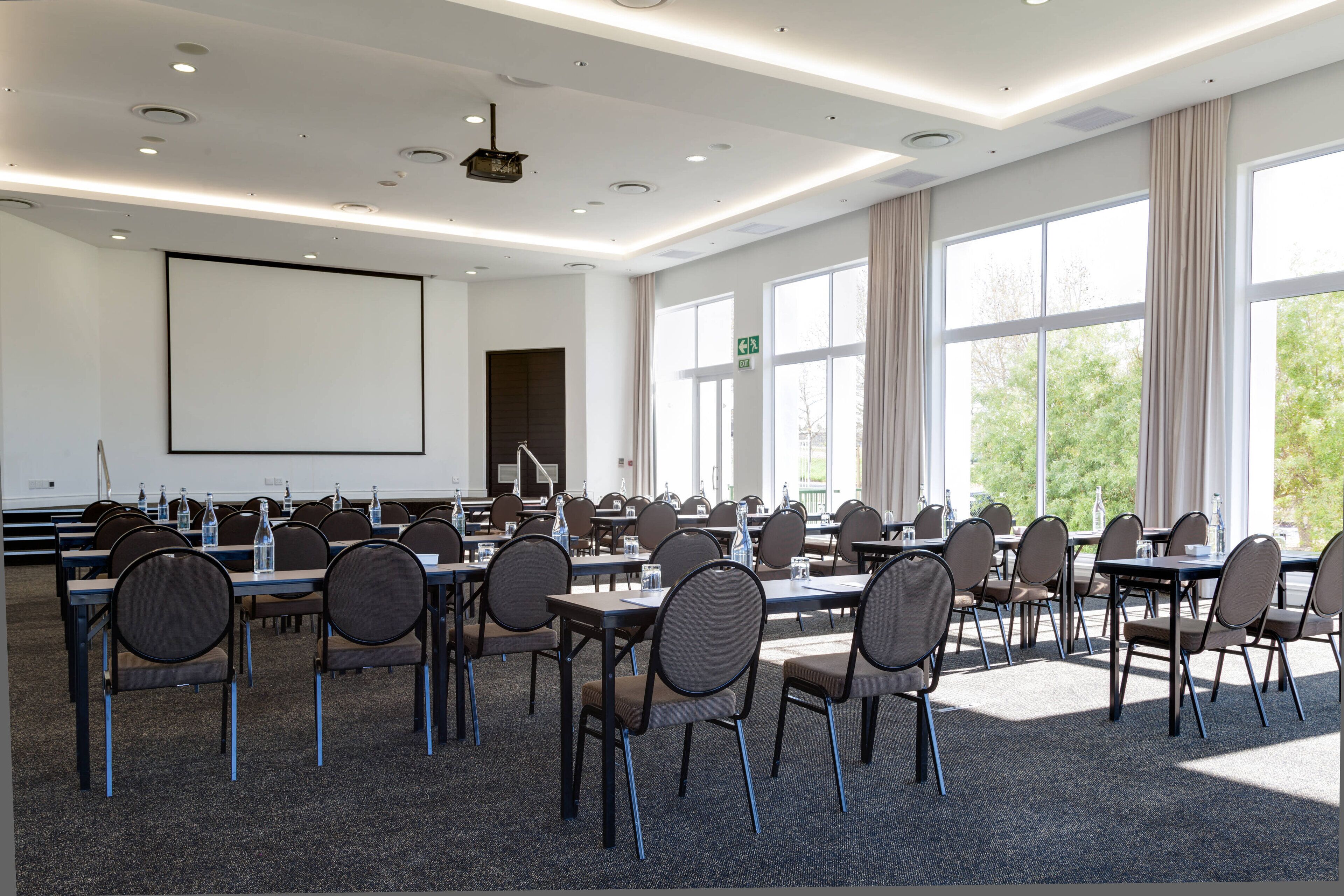 Une salle de conférence dotée de plusieurs tables et chaises, de grandes fenêtres et d’un écran de projection.