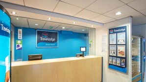 L’espace d’accueil d’un hôtel Travelodge à l’aéroport de Glasgow, avec un comptoir d’accueil en bois, un écran numérique et une vitrine contenant des brochures.