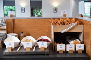 A breakfast buffet with labeled bowls of natural yogurt, granola, and fruit compote, and a wooden display of muffins and a floured bap.