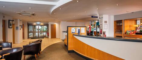 Un hall de hotel con mostrador de recepción, zona de estar y señalización para la zona de bebidas.