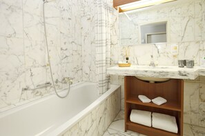 A bathroom with a marble-tiled wall, a bathtub, a wooden vanity with a sink, and a mirror. - Hotel Steinmattli (Adelboden)