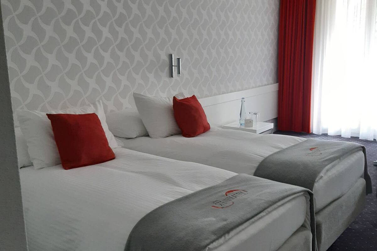 A hotel room with two beds, a patterned wall, a red curtain, and a nightstand with a lamp.