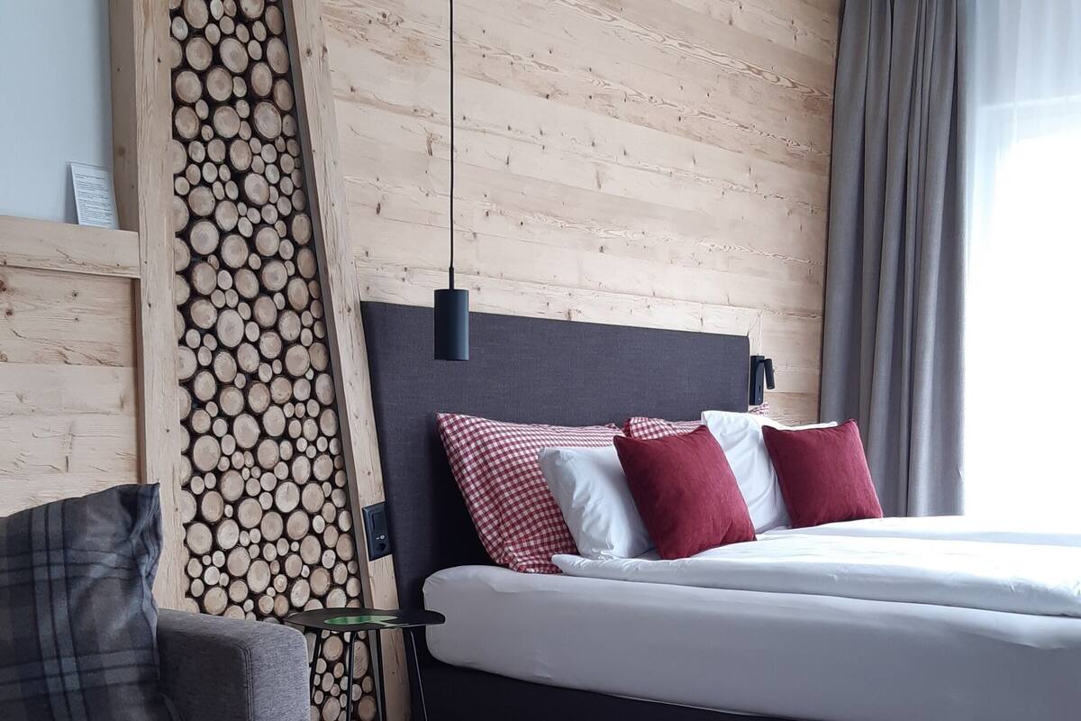 A hotel room with a wooden wall, a bed with red and white pillows, a grey sofa, and a round wooden table with a clear bottle and glasses.