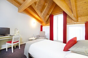A hotel room with a wooden ceiling, a bed with red and grey pillows, a desk with a computer and chair, and a window with red curtains. - Hotel Steinmattli (Adelboden)