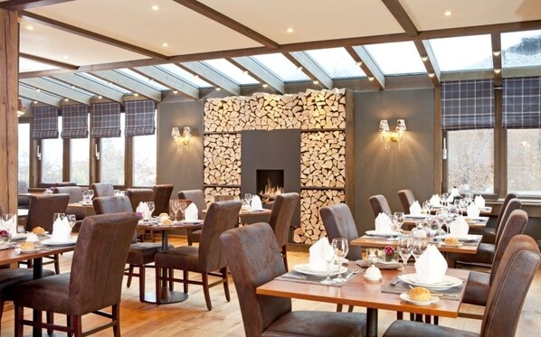 Un restaurante con pared de piedra y chimenea, mesas y sillas de madera, y ventanas amplias.