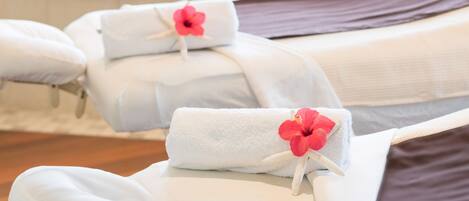 A spa room with two massage tables, each with a towel and a red flower. A bathtub with a faucet and a plant is visible in the background.