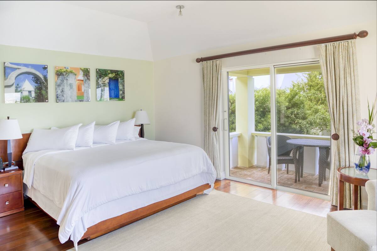 A bedroom with a large bed, a nightstand, a lamp, and a view of a balcony with a table and chairs.
