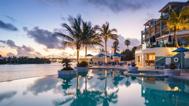 Zona de piscina en un resort con sillones de playa y sombrillas, un edificio grande con balcones y vistas al agua y al horizonte de la ciudad al atardecer.