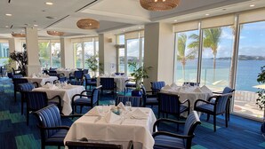 Un restaurante con mesas dispuestas para comer, amplias ventanas con vista al mar y sillas a rayas azules.