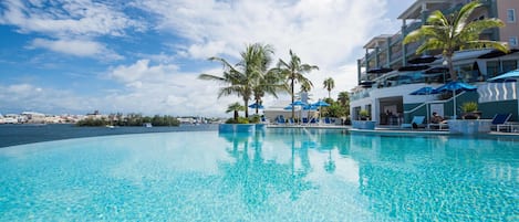 A clear blue pool with lounge chairs and umbrellas, surrounded by palm trees and a building with balconies.
