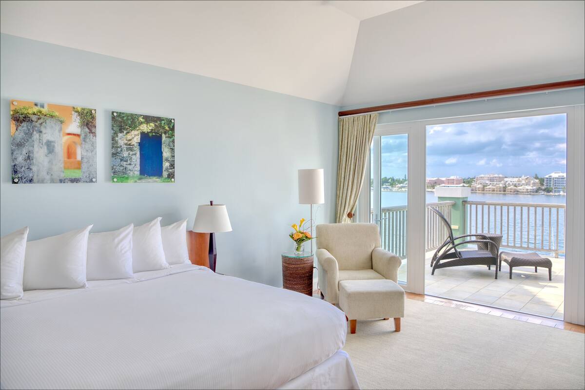 A hotel room with a large bed, a chair, a table with a vase of flowers, and a balcony with a view of the sea.