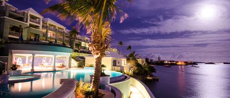 A resort with a swimming pool, palm trees, and a view of the water at night.