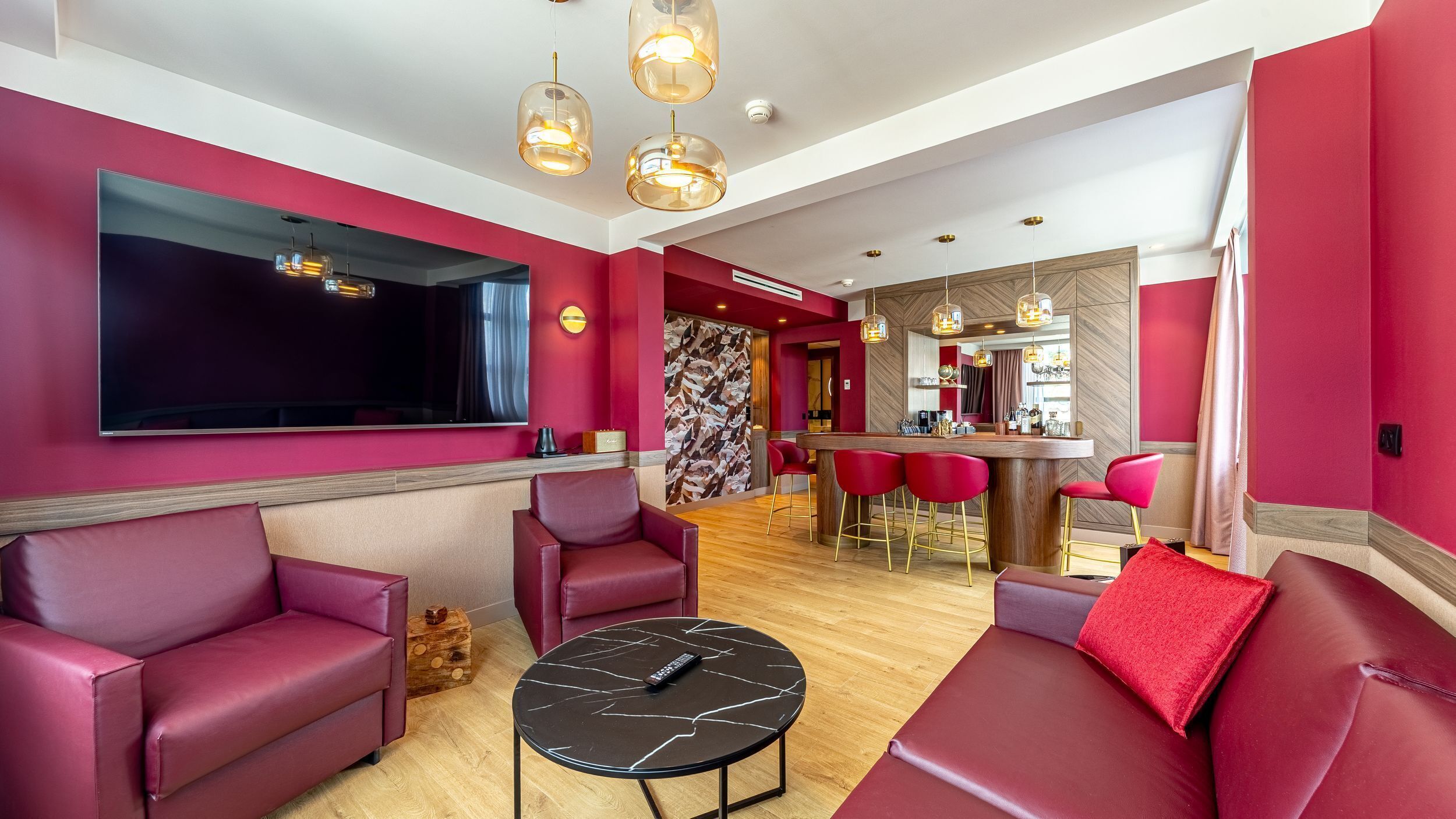A modern living room with red sofas, a large flat-screen TV, and a bar area with stools.