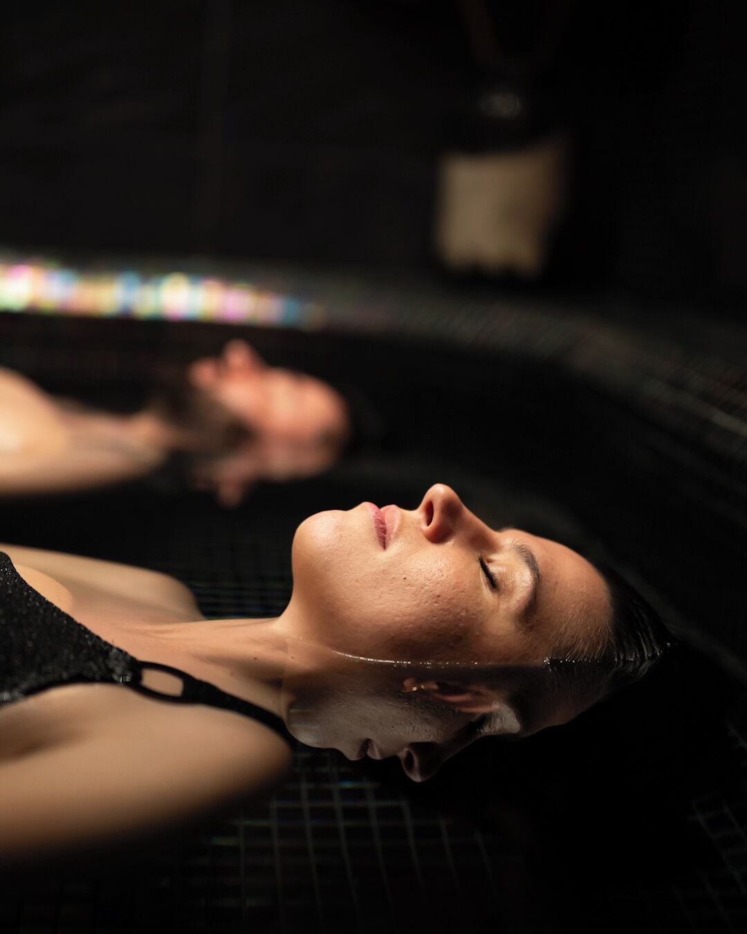 Une personne allongée dans une pièce sombre, dont le sol est recouvert d’un motif quadrillé, peut-être dans un espace de soins ou de détente.