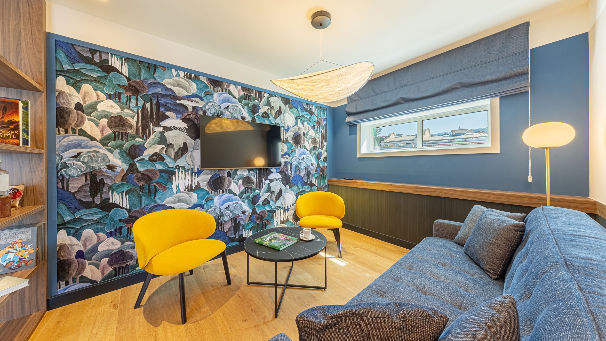 A modern living room with a blue wall, a large mural, a sofa, two yellow chairs, a coffee table, and a TV.