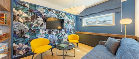 A modern living room with a blue wall, a large mural, a sofa, two yellow chairs, a coffee table, and a TV.