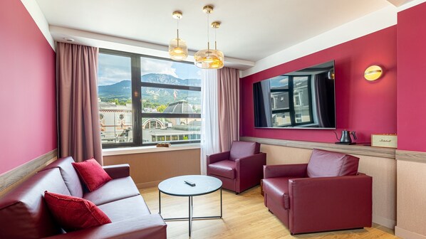 A modern hotel room with a red and beige color scheme, featuring a sofa, armchairs, a small table, and a large window offering a city view.