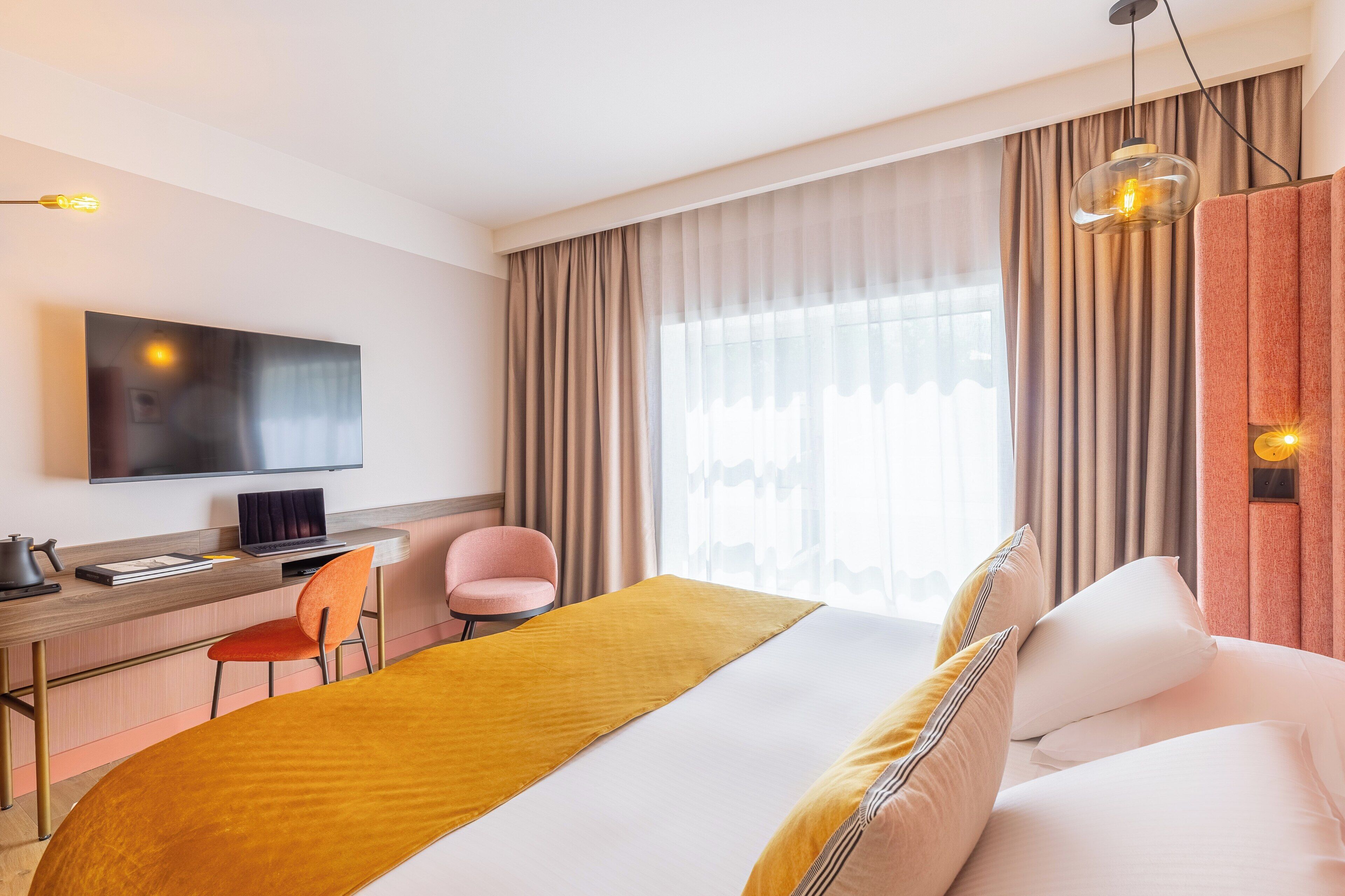 Ein Hotelzimmer mit Bett, Schreibtisch, Laptop, Stuhl, Fernseher und Vorhängen.