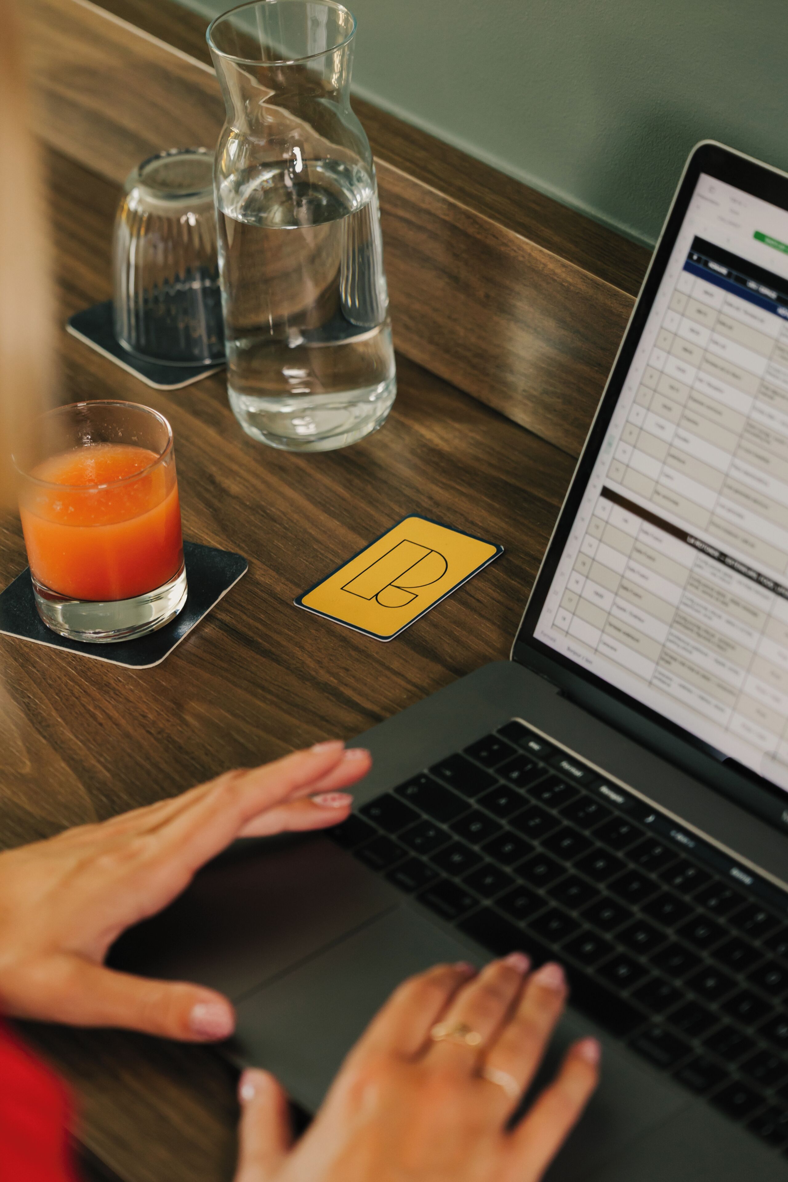 A laptop on a wooden table with a glass of orange juice, a water bottle, and a sticky note with a drawing.