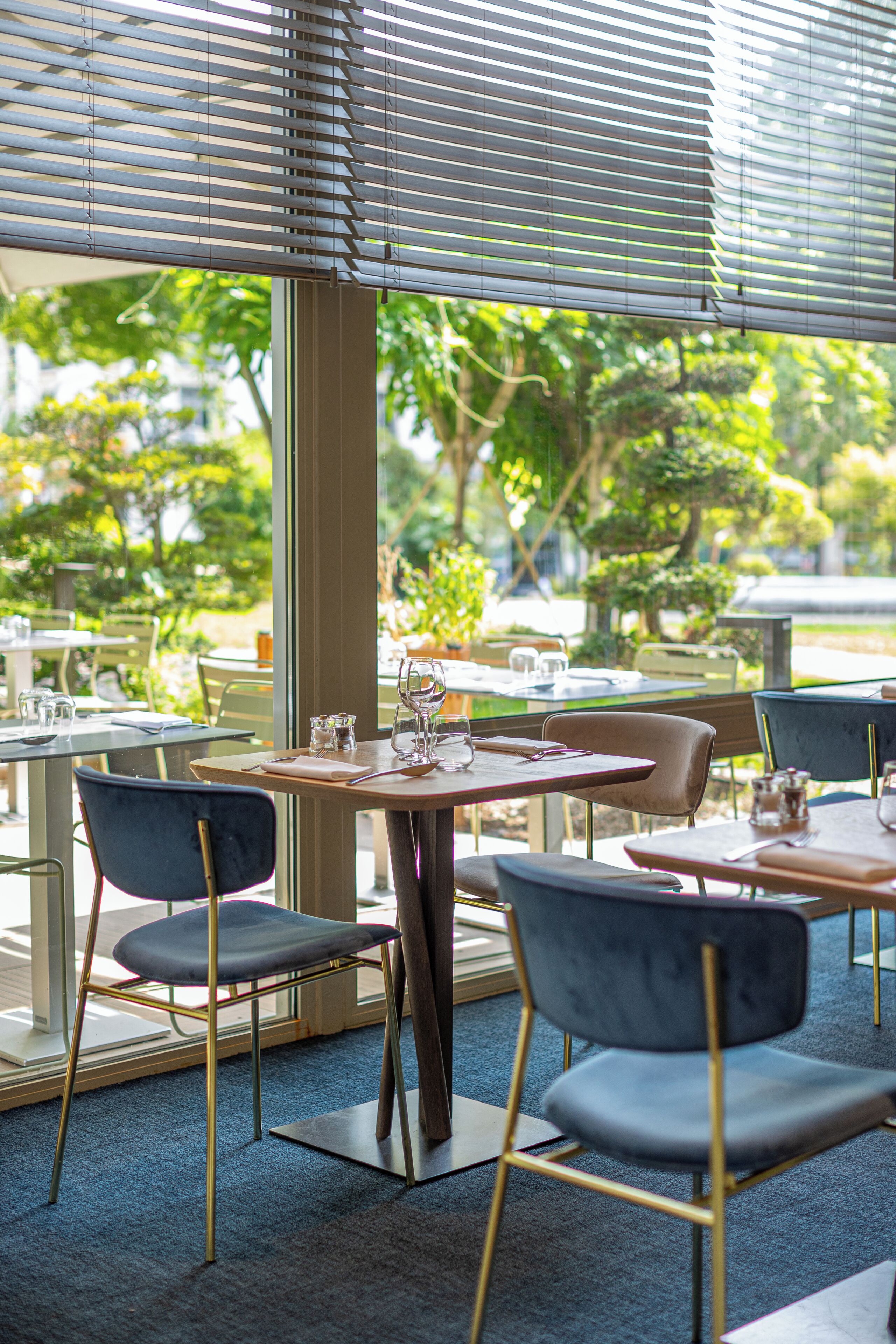 An outdoor dining area with tables and chairs, a view of greenery, and a building with large windows.