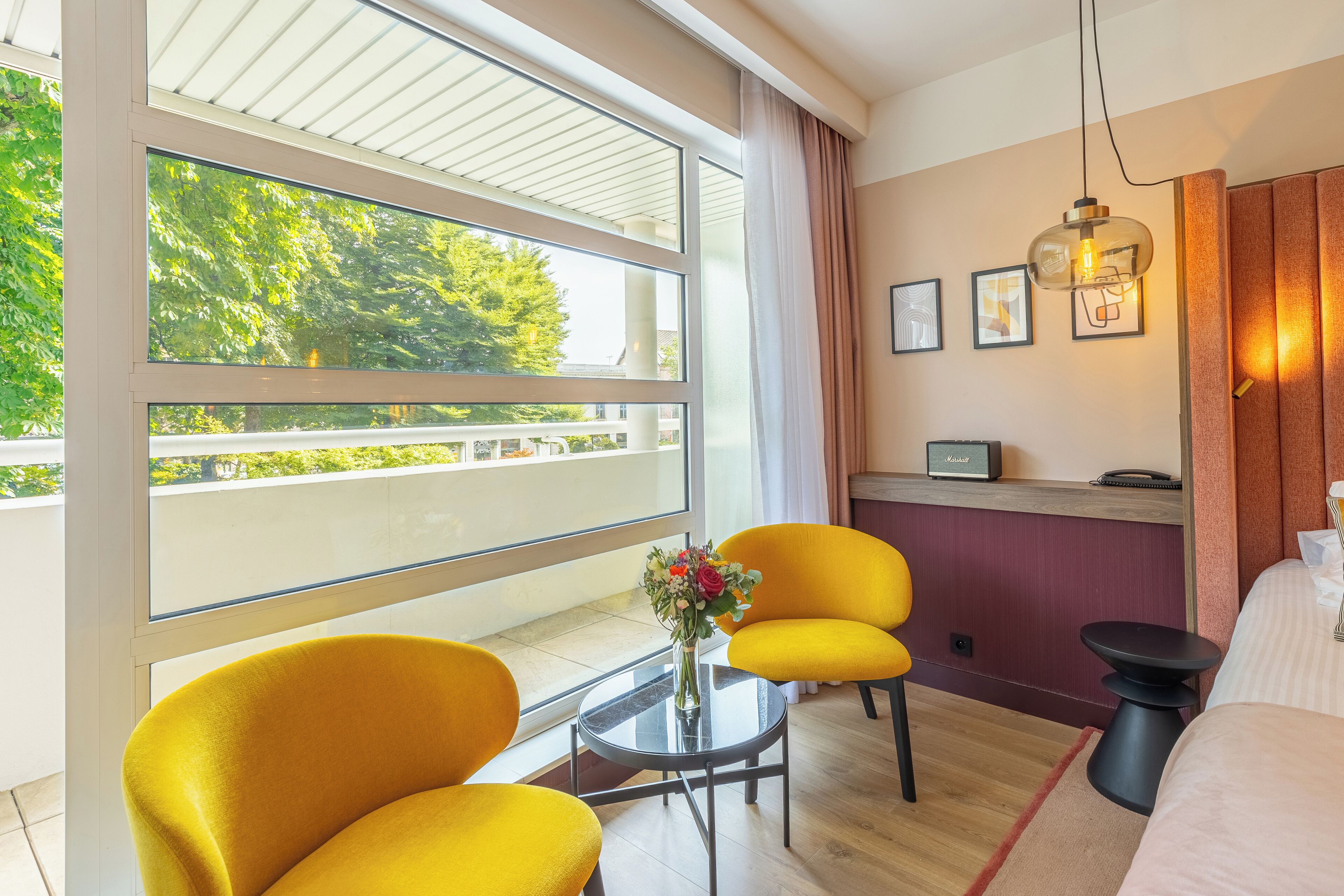 A hotel room with a large window, a small table with a vase of flowers, two yellow chairs, a bed, and a desk with a computer.