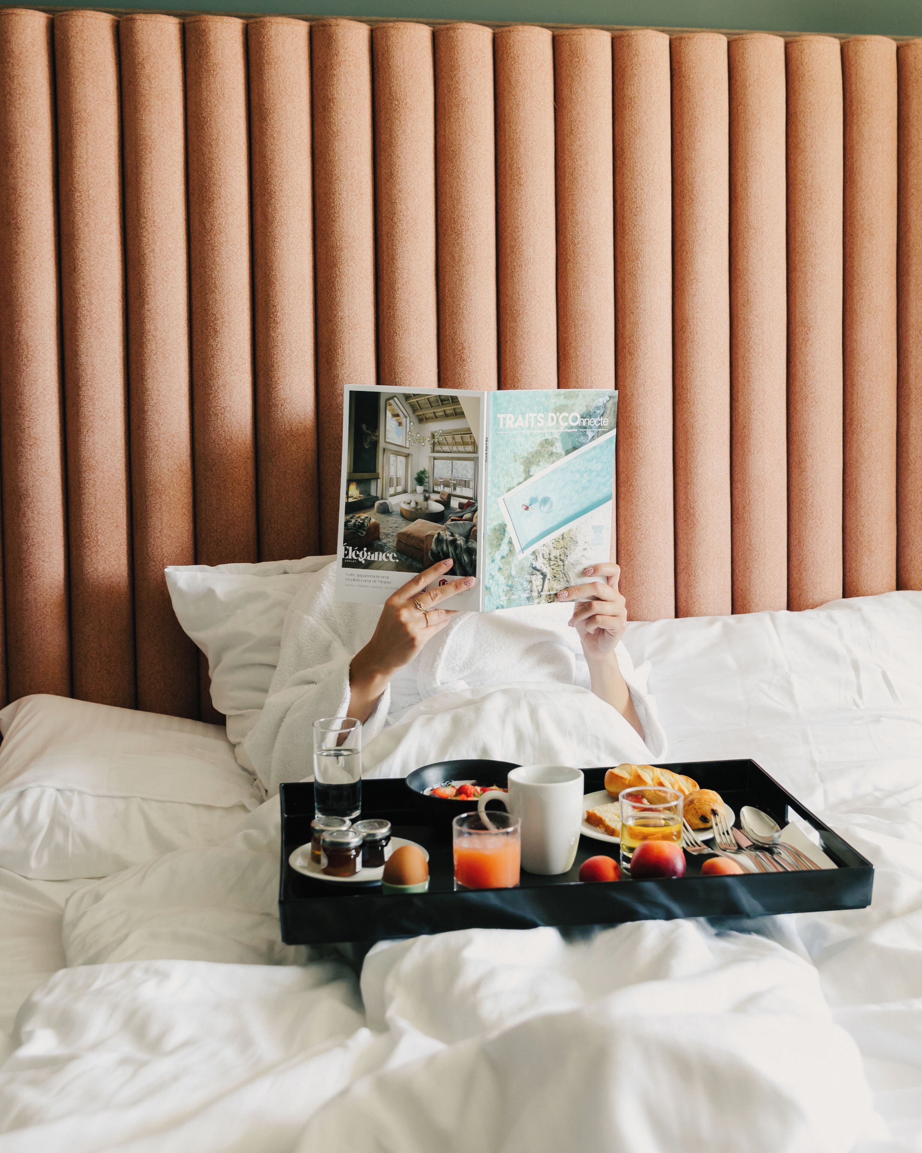 Een persoon ligt in bed, een ontbijttafel met spullen ernaast, leest een tijdschrift.