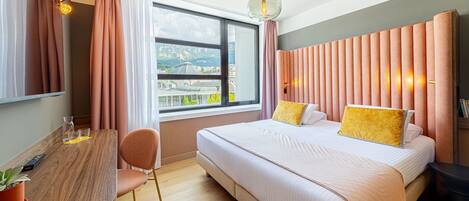 Moderni hotellihuone, jossa on suuri sänky, puinen työpöytä, tuoli ja ikkunasta avautuva näkymä kaupunkiin.
