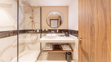 Un baño moderno con ducha envuelta en vidrio, espejo redondo y tocador de madera con lavamanos.