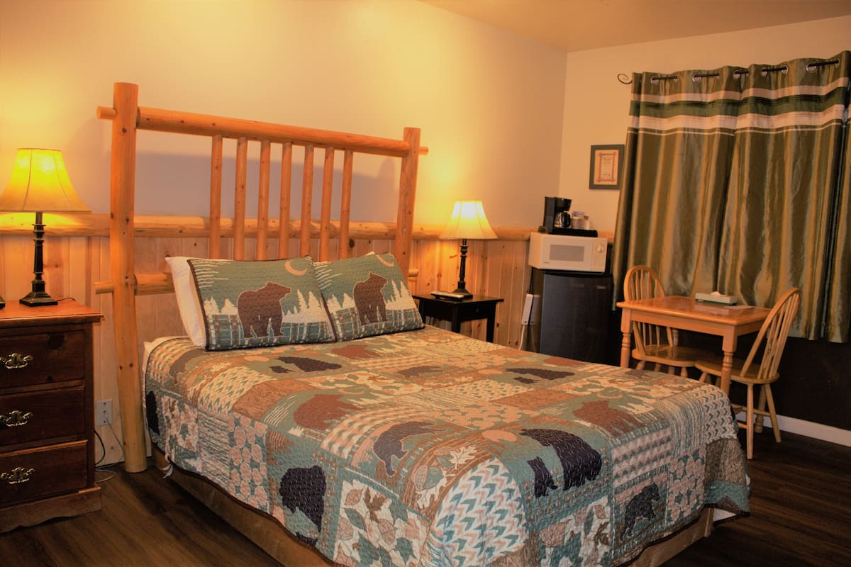 Ein Schlafzimmer mit einem hölzernen Bett, zwei Nachttischlampen, einem Schreibtisch mit Stuhl, einer Mikrowelle und einem Vorhang.