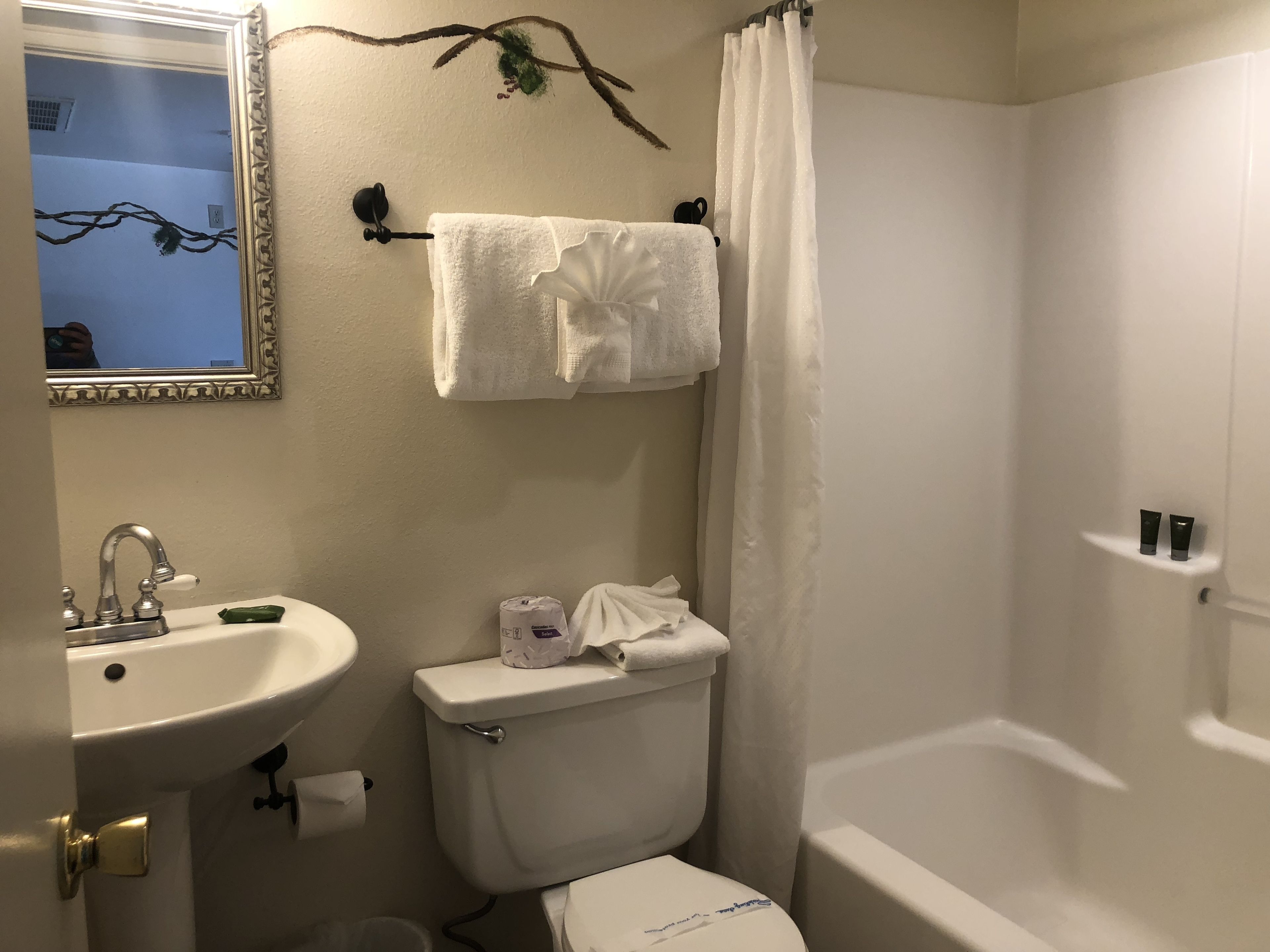 Un baño con lavamanos, inodoro y bañera. Hay un perchero con una toalla, un rollo de papel higiénico y un pequeño estante con artículos de aseo.
