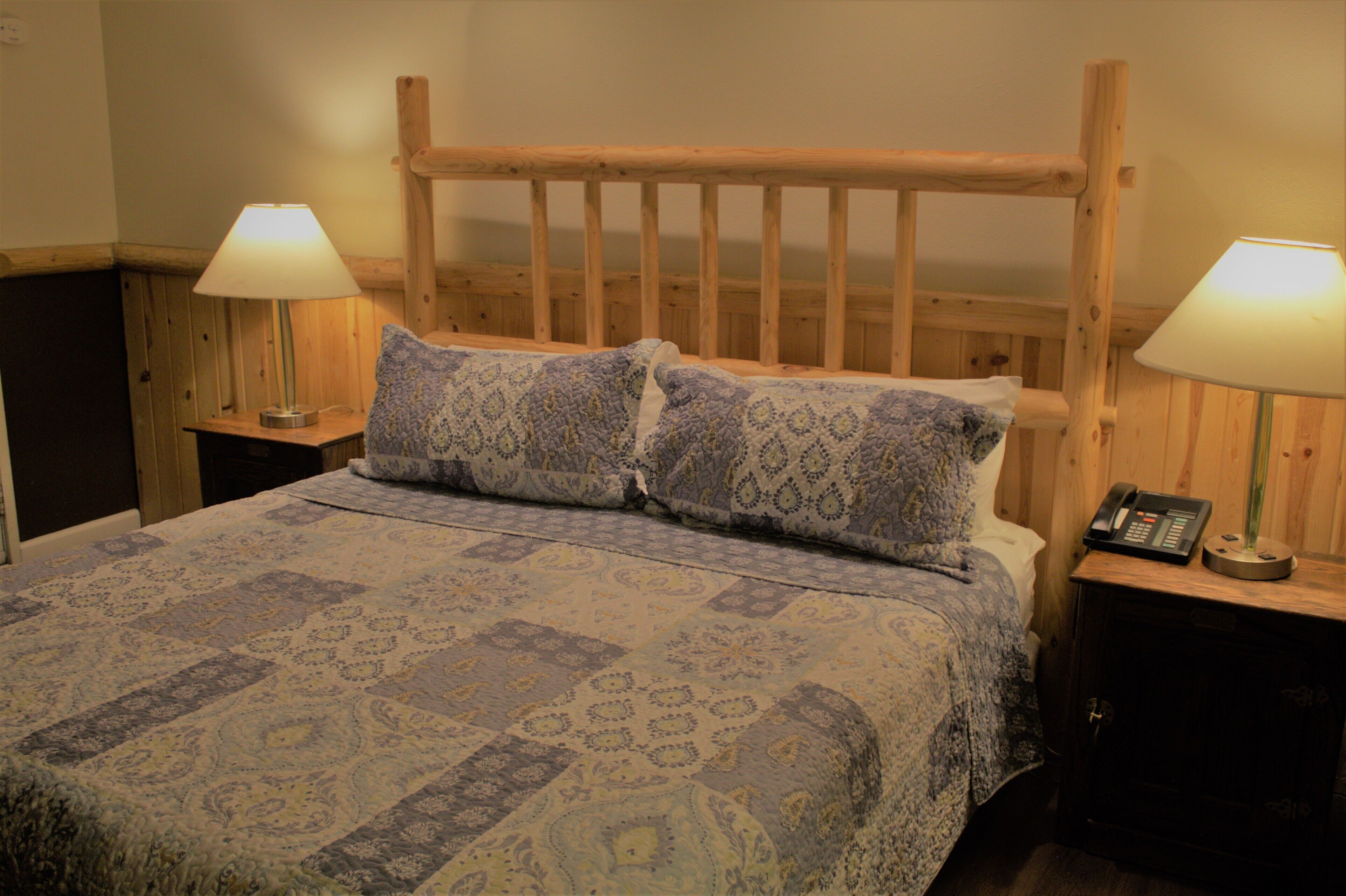 A bed with a patterned bedspread, two pillows, wooden headboard, bedside lamps, and a nightstand with a phone.