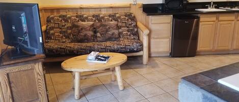 Una sala de estar con un sofá estampado, una mesa de centro, un televisor sobre un mueble de madera, y una zona de cocina con armarios y un fregadero.