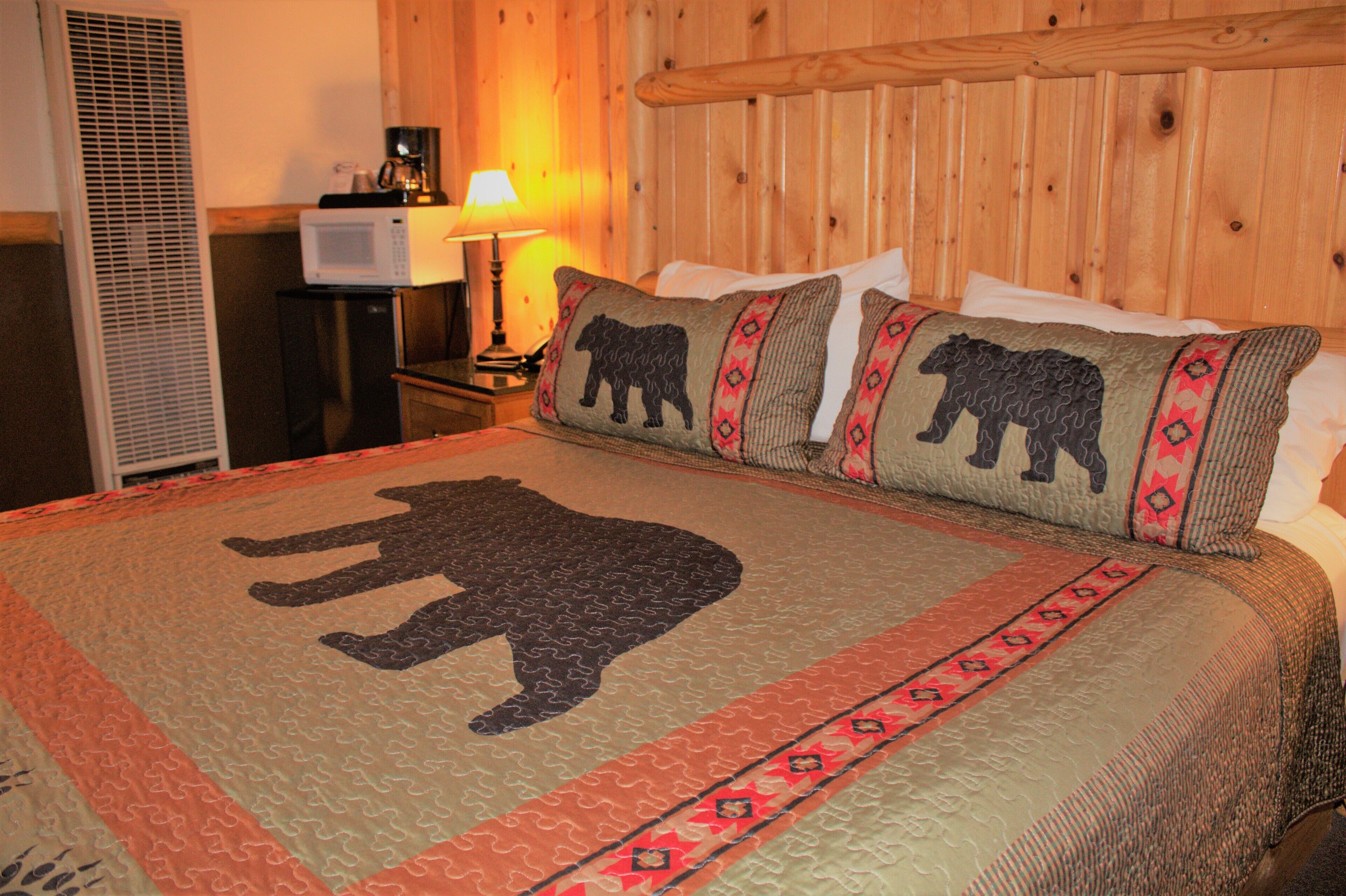 A hotel room with a bed featuring a bear-themed quilt, a wooden headboard, a nightstand with a lamp, and a small television.