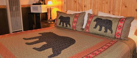 A hotel room with a bed featuring a bear-themed quilt, a wooden headboard, a nightstand with a lamp, and a small television.