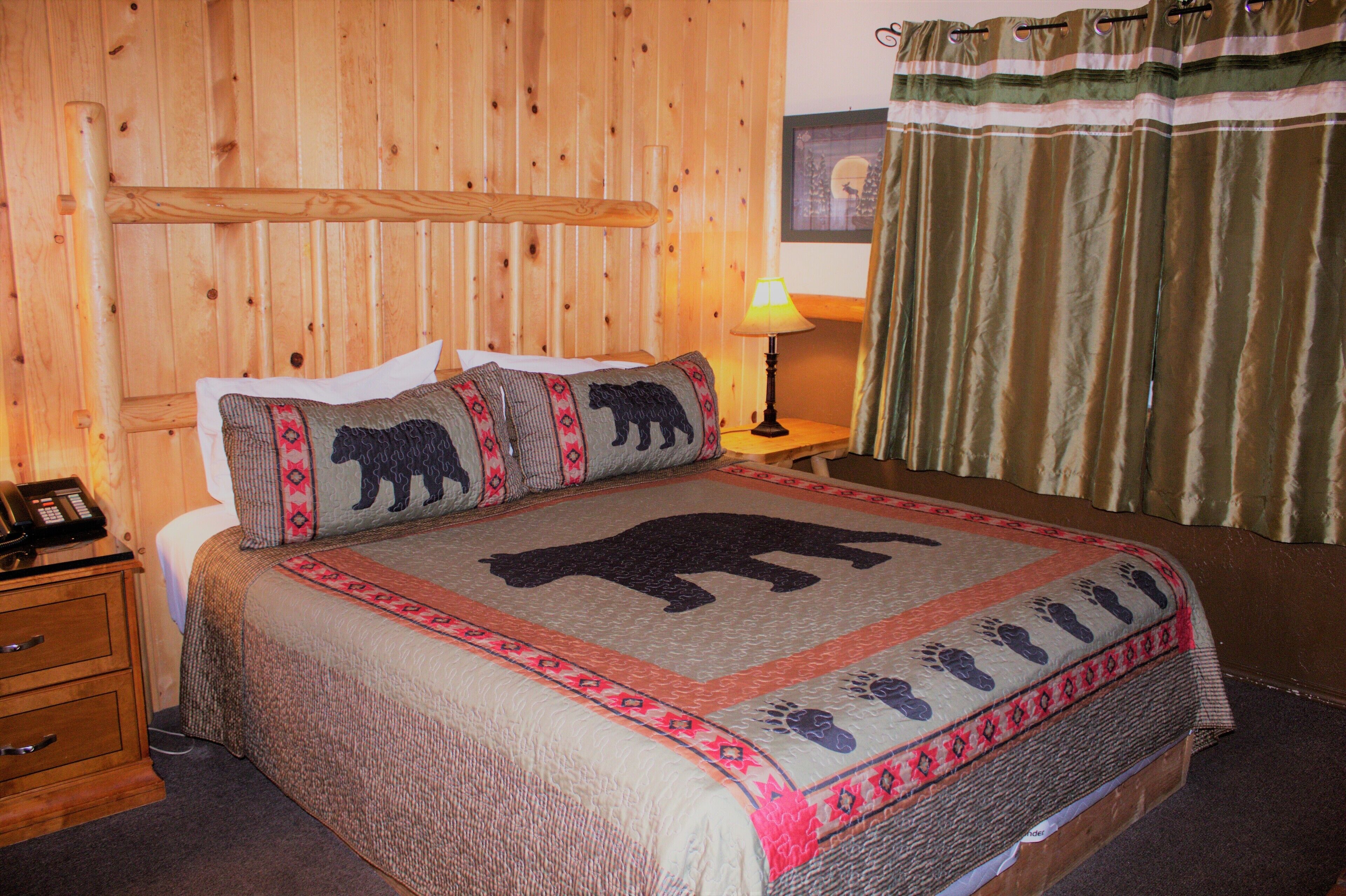 Habitación con paneles de madera, una cama con edredón con estampado de oso, una lámpara de noche y un teléfono sobre una mesita de noche de madera.