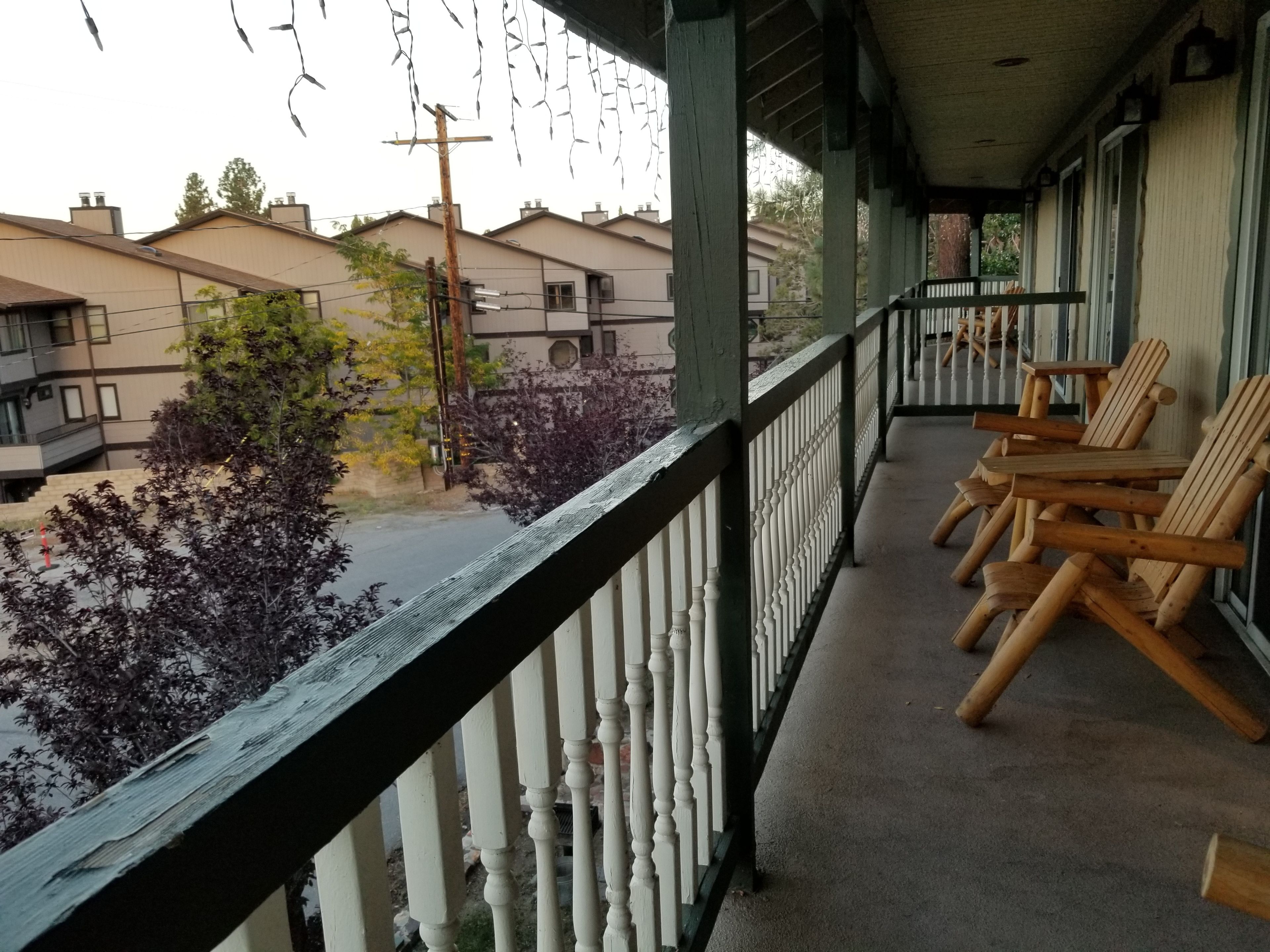 Een balkon met houten stoelen en een witte balustrade met uitzicht op een straat met huizen en bomen.