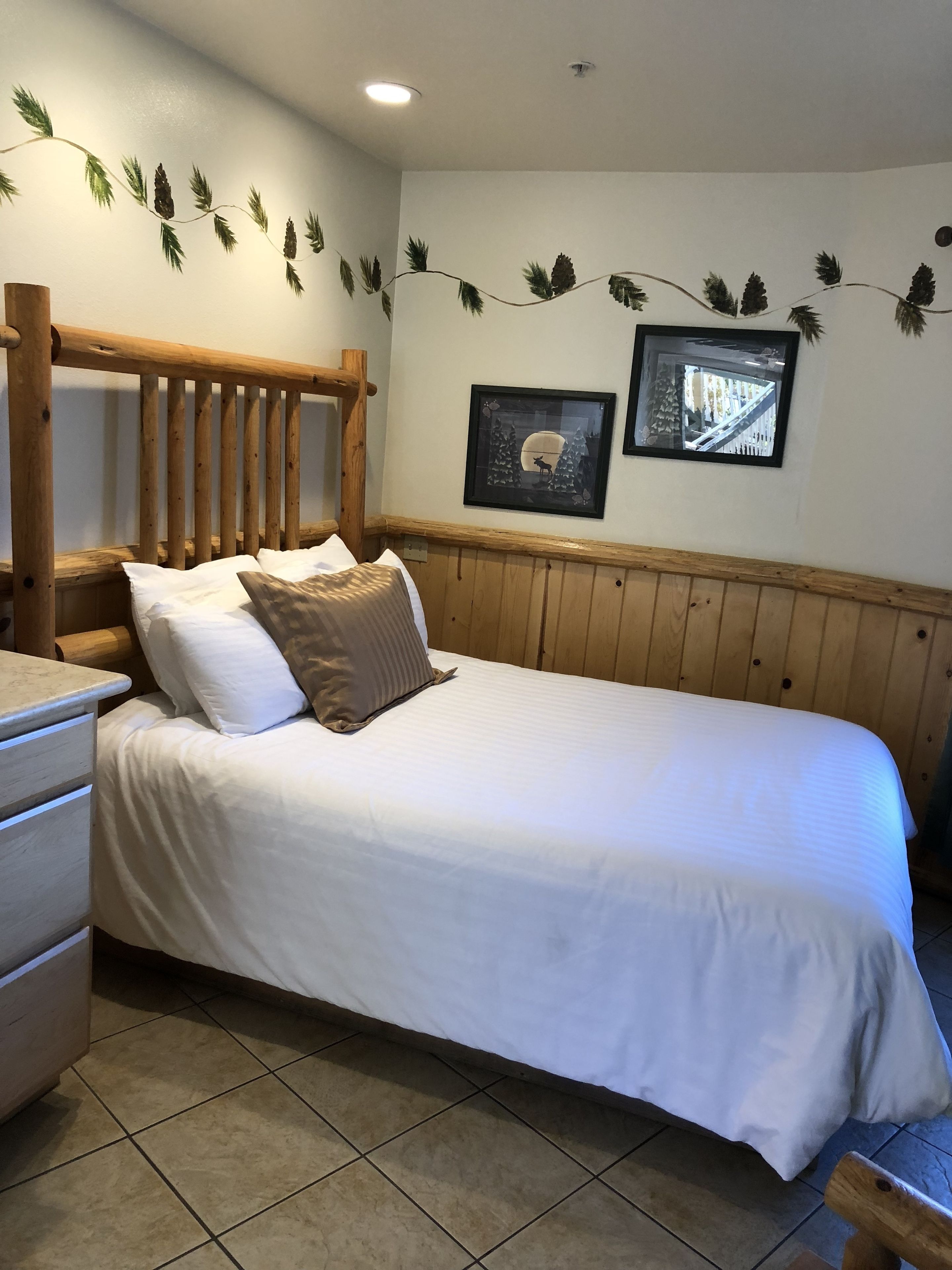 Un lit bien fait, avec une literie blanche et une tête de lit en bois, une commode en bois avec un miroir, et deux tableaux encadrés accrochés au mur.