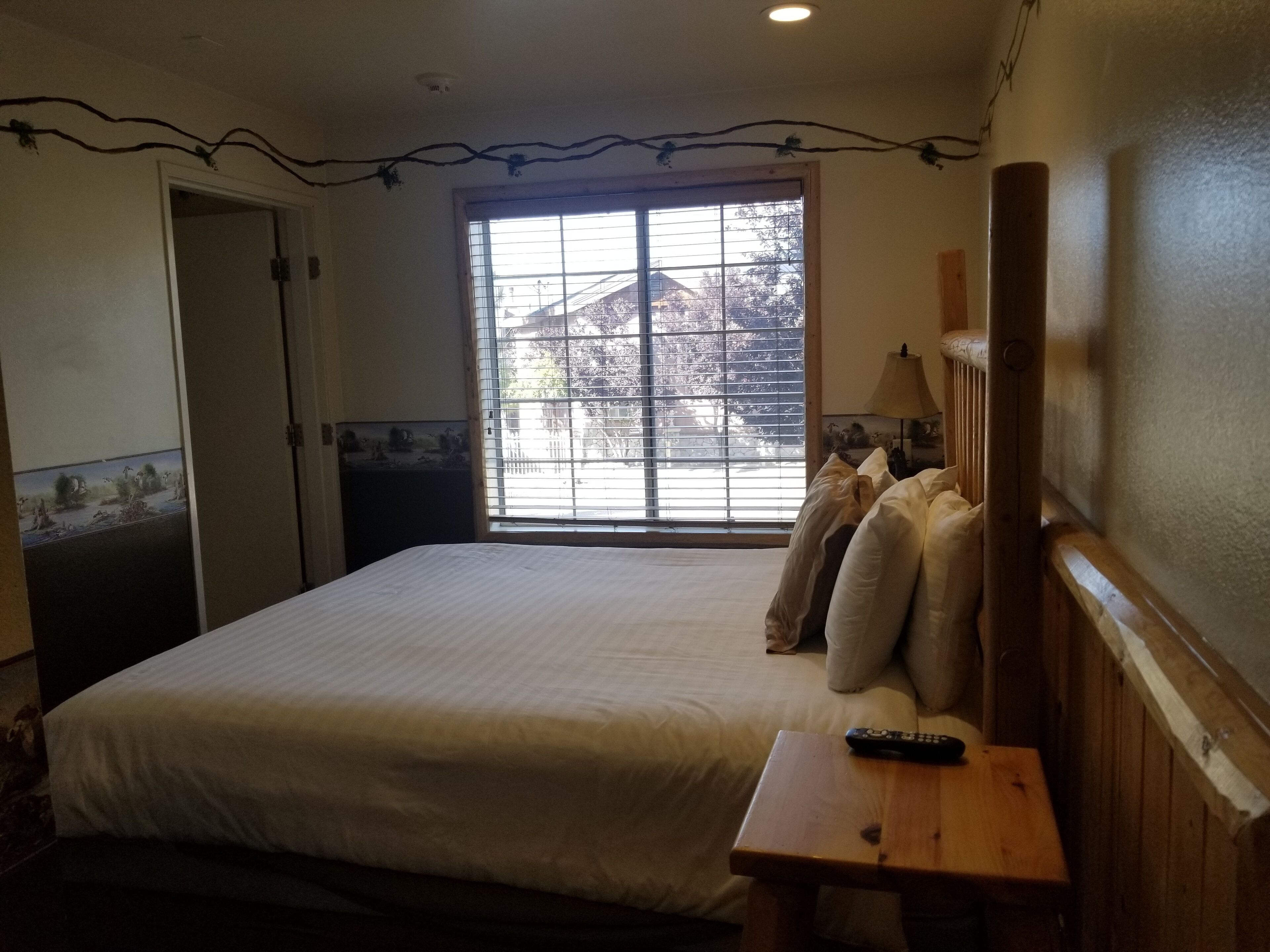 A bedroom with a large bed, a wooden headboard, a window with blinds, a bedside table, and a remote control.