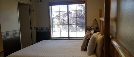 A bedroom with a large bed, a wooden headboard, a window with blinds, a bedside table, and a remote control.