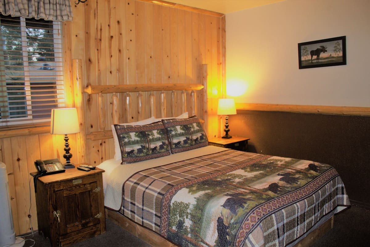 Ein Zimmer mit Holzwandverkleidung, einem Bett, zwei Nachttischlampen, einem Nachttisch mit Telefon und einem gerahmten Bild an der Wand.
