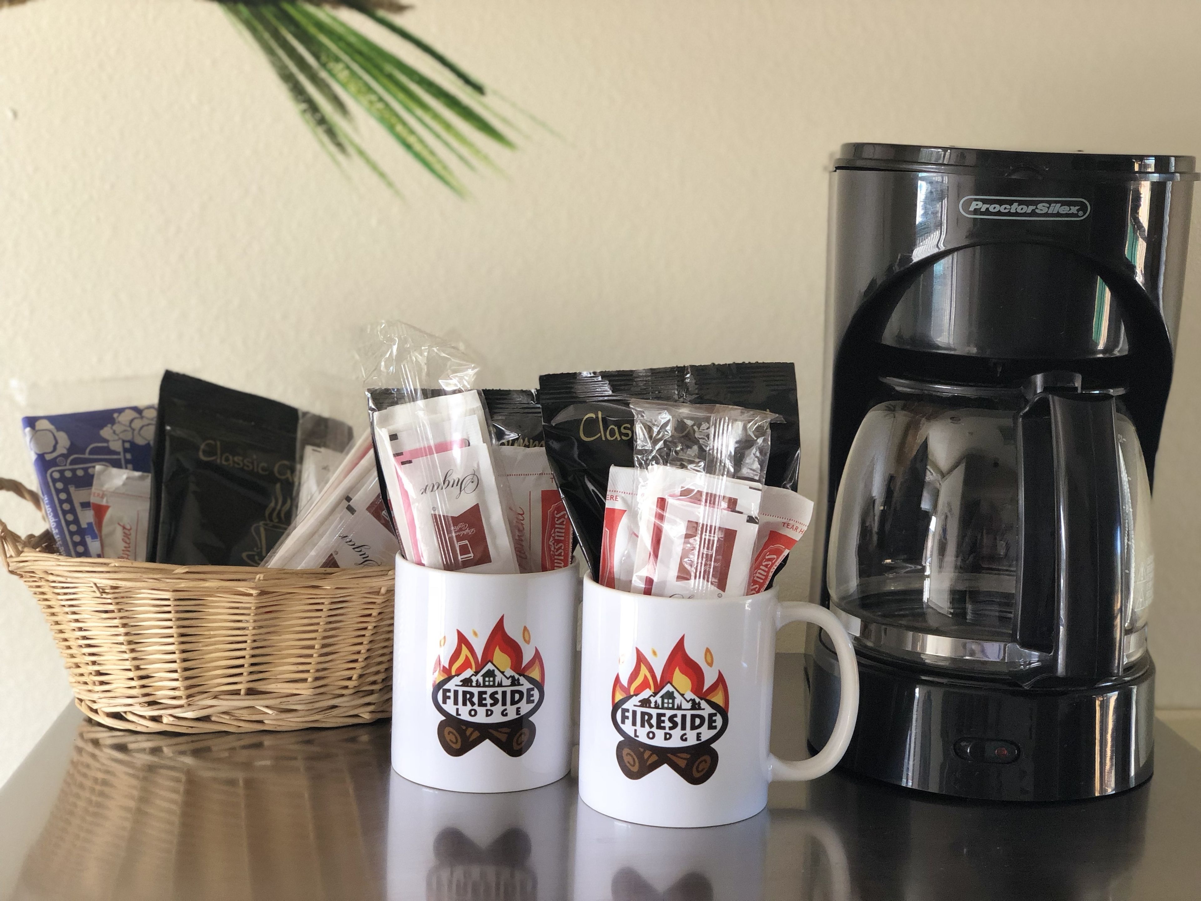 Dos tazas blancas con el logo de "Fireside Lodge", una cafetera Proctor Silex y una cesta de mimbre con varios objetos.
