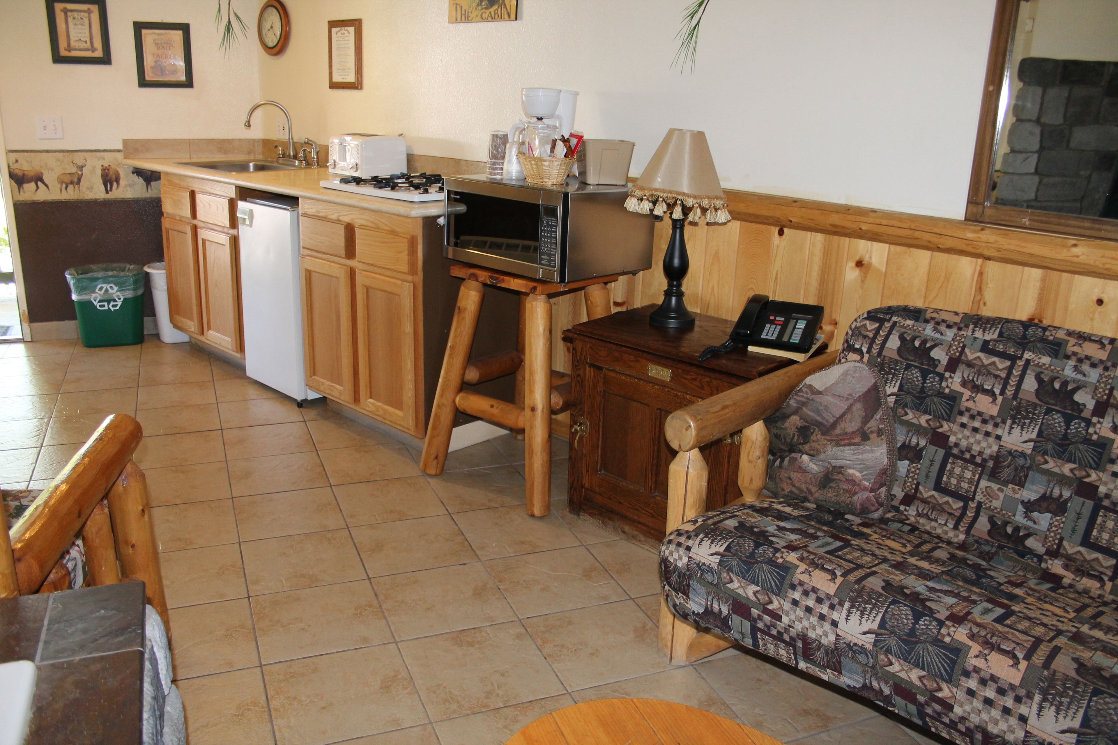 A compact kitchen with a microwave, sink, and wooden cabinets. There is a patterned sofa and a small table with a lamp.