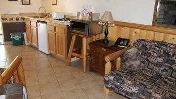 A compact kitchen with a microwave, sink, and wooden cabinets. There is a patterned sofa and a small table with a lamp.