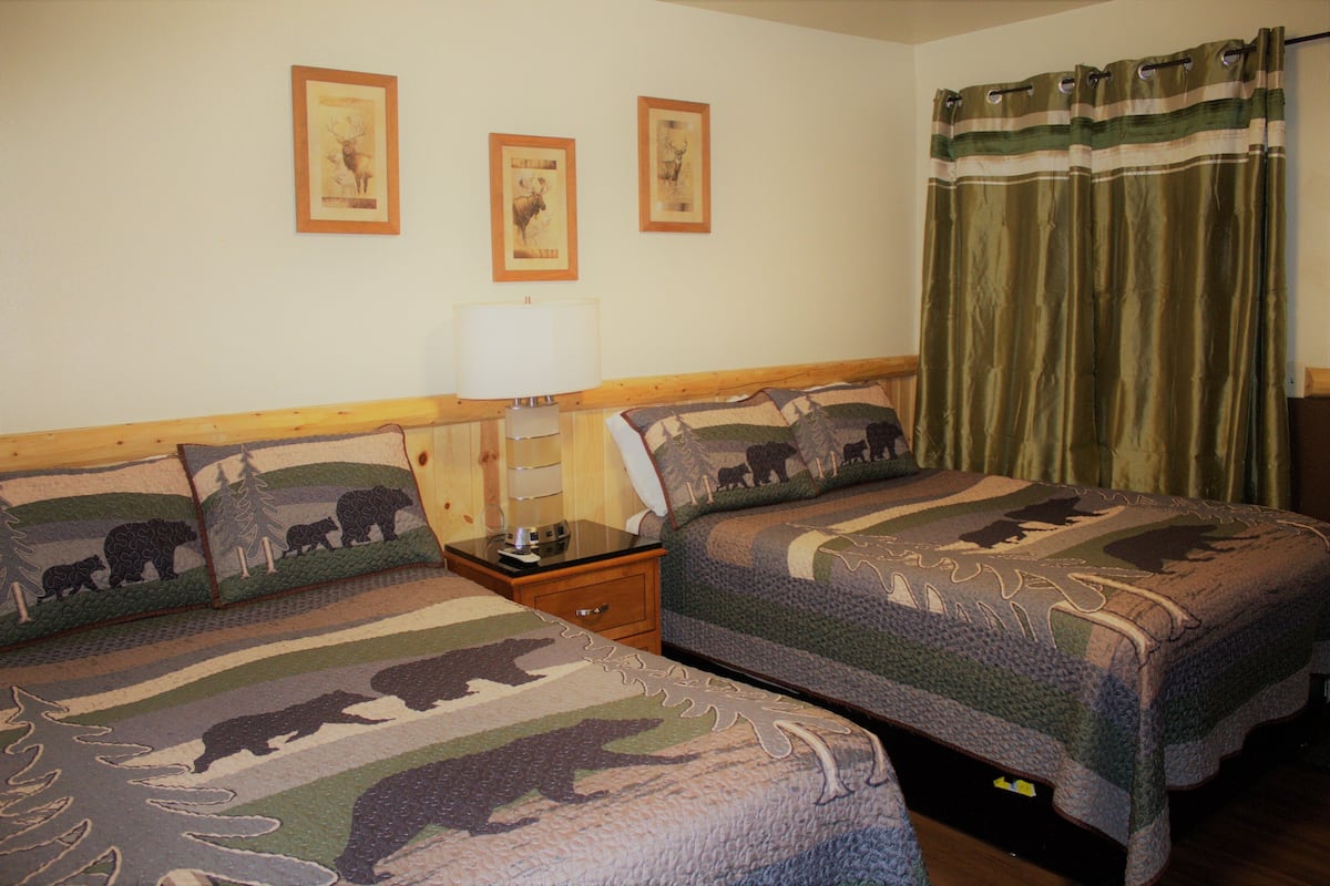Ein Zimmer mit zwei Betten, jeweils mit einer bettenbezogenen Decke mit Bärenmotiv, einem Nachttisch mit Lampe und einem Fenster mit grünem Vorhang.